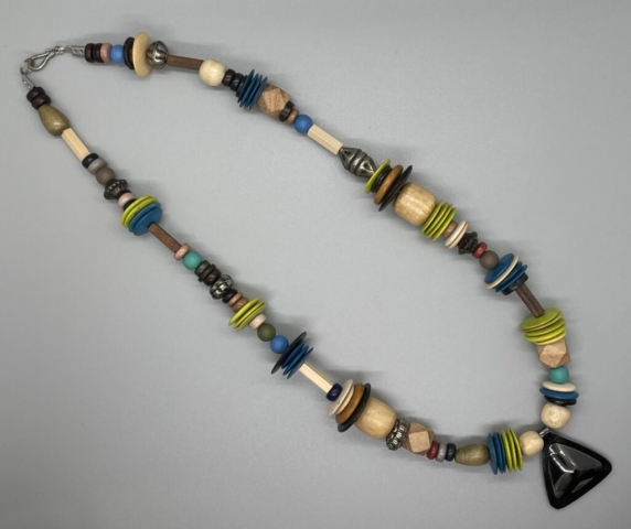 Unisex Neckwear- Green Tear Shaped Wood Beads. Multi-Shape Pewter Ribbed Beads. 4 Sided Beige and Brown Wood Beads. Natural Barrel Wood Beads. Multi-Color Coconut Shell Discs. Blue, Green and Turquoise Round Glass Beads. Faceted Wood Beads. Multi-Color Earth Tone Rondelle Beads. Small Natural Wood Beads. 1 1/4 inch Gunmetal-Plated Pewter Single-Sided Triangle Centerpiece. Approx. length: 23 inches. Pronounced: GO-WAHN. Origin: Africa. Meaning: Rainmaker