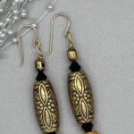 Earrings- Embossed, Brass Metalized Acrylic Old World Tube Bead. Black Opaque Czech Crystal Bicone Beads. Orange Heishi Shell Beads. Pewter Textured Barrel Beads. Approx. length: 2 1/2 inches. Hand Shaped 14K Gold Filled Earwire. Pronounced: JAH-HA. Origin: Africa. Meaning: Dignity