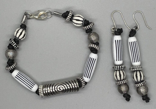 Wrist Set- White Old World Long Tube, Round and Disc Beads. Pewter Round Beehive Beads. Silver Metal Tube Bead with Etched Swirl Design. Knotted Design done with Black Nylon Cord. Approx. length: 8 inches. Hand Shaped .925 Sterling Silver Earwire. Approx. length: 3 inches. Pronounced: N-DOO-LOO. Origin: Africa. Meaning: Dove
