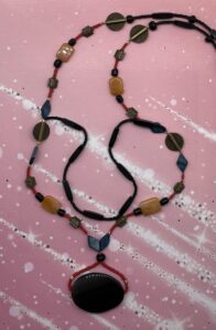 Necklace- Black and Red Acrylic Pony Beads. Round and Barrel Shaped Glass Beads. Acrylic Four Sided Tube Beads. Gold Metal Carved Discs. Square Carved Pewter Beads. Blue Mother-of-Pearl Beads. Rectangle Kyanite Beads. Natural Stone Black Onyx Centerpiece. Approx. length: 29 3/4 inches. Attachment length: 10 inches. Pronounced: em-CHUM-ba. Origin: Eastern Africa. Meaning: Sweetheart